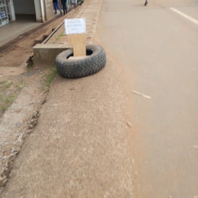 The scene shows a dirt and paved roadway in Sheywe ward, Kenya. A "STRICTLY NO PARKING HERE" sign is propped up by a tire on the edge of the road near a sidewalk. Several people are walking in the background.