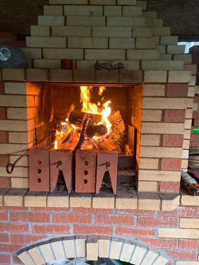The image depicts a brick fireplace with a fire burning inside. The fireplace is constructed of light-colored and reddish-brown bricks in a patterned design. Two brick structures with triangular notches are positioned inside the fireplace. A metal poker rests near the base of the fire. The location is Gryaznoye, Russia.