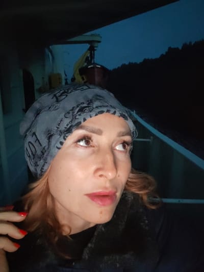 A woman wearing a gray bandana with black lettering and a dark fur-lined jacket is pictured outdoors at night. She has blonde hair and is looking up and to her left. Her fingernails are painted red. In the background, a dark silhouette of trees and a faint blue sky are visible, suggesting an outdoor location. There are no discernible city landmarks to indicate Annenskii Most, Russia.