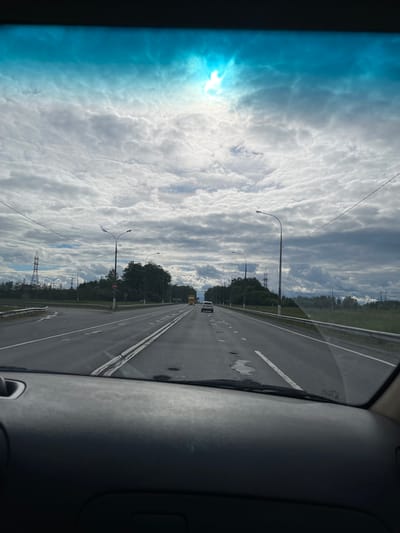 The image shows a view from inside a vehicle moving along a highway. The road is straight, divided by double white lines, and has streetlights along the side. There's a cloudy sky with the sun shining through, visible on the left. Vegetation is present on the roadside, with power lines and towers visible in the distance. The dashboard of the vehicle is visible on the right side of the image. The location is Starokurovo, Russia. There are no people visible.