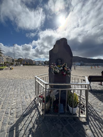 The image depicts a monument on a cobblestone promenade overlooking a sandy beach in Chayofita, Spain. The monument is a large, dark, irregular-shaped stone with a metal plaque and a floral arrangement at its base, all enclosed by a silver metal railing. The flowers include a mix of roses and orchids in various colors. Inside the railing, a potted snake plant and other small potted plants are visible.

Beyond the monument, the beach stretches out, dotted with people. Some individuals are playing volleyball, while others are walking along the shore. The ocean is calm, and in the background, a line of multi-story buildings and palm trees forms the cityscape. Mountains rise in the distance.

The sky is partly cloudy with bright sunshine, casting long shadows from the monument and railing onto the cobblestones. The overall scene suggests a sunny day in a coastal tourist area.