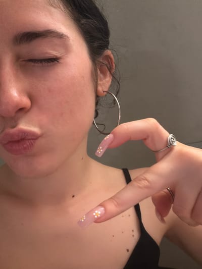 The image shows a close-up of a person's face, laying down with eyes closed. The person has dark hair and light skin. They are wearing a silver hoop earring and have a hand raised above their face. The hand has long, light pink nails decorated with small white flowers. A silver ring is on the raised hand. Part of a black garment is visible on the shoulder area. The background is a solid light grey. It is not possible to determine the location is Lissone, Italy, from this image.