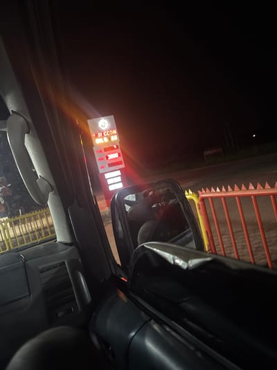 The image is taken from inside a vehicle, likely a car, at night. The driver's side mirror is prominently visible, reflecting a person and a light source. Outside the vehicle, a fuel station sign is illuminated, displaying prices including "2000". A barrier or fence is positioned nearby on the right side of the image. Based on the sign visible in the photo, the location is likely Ozalla, Nigeria.