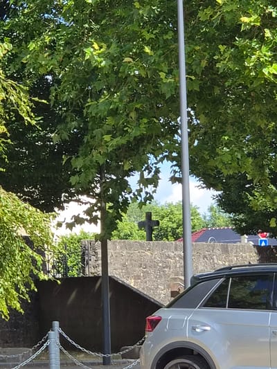 This is a vertical, eye-level, outdoor shot of a stone wall and lush green trees. A portion of a silver vehicle is visible on the right side of the frame, parked next to the stone structure. The vehicle appears to be a van or SUV, with its rear taillight and part of a wheel showing. A metal pole runs horizontally across the middle of the image, in front of the trees and wall.
Further back, a stone cross is visible, set against a backdrop of trees and a bright sky, suggesting it might be a cemetery or a religious site. There is a hint of a blue sign with a white arrow and red accents on a pole in the distance, indicating a road or direction. The overall scene suggests a sunny day, with dappled light filtering through the leaves of the trees. The image is captured in Schengen, Luxembourg. No people are visible in the frame.