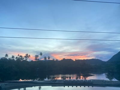 This is a photograph of a tropical landscape at dusk, likely in Lambaro Kueh, Indonesia. The sky is a gradient of blues and purples, with streaks of orange and pink indicating the setting sun behind a silhouetted mountain range. A line of palm trees stands against the colorful sky, their fronds casting dark shadows. Below the trees, a body of water reflects the twilight hues and the shapes of the vegetation. In the foreground, a concrete structure with multiple openings runs horizontally across the bottom of the frame, its surface also reflecting the scene above. Power lines are visible stretching across the upper portion of the image. The overall impression is one of natural beauty and tranquility at the end of the day.