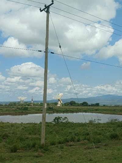 A wooden utility pole stands in a grassy field in Maai Mahiu, Kenya. It is holding up a set of power lines that run across the sky. There are two satellite dishes visible in the background, along with a small body of water that appears to be a pond. The sky is blue with fluffy white clouds. The surrounding land is mostly green and appears to be a mix of grass and brush. The coordinates provided are near the Athi River and the Nairobi-Nakuru highway. The image does not depict any people or actions.