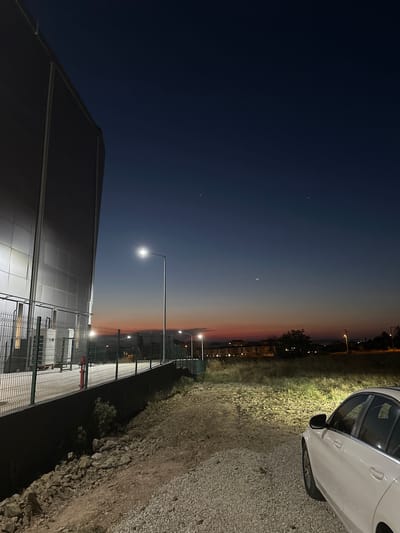 The image depicts a twilight scene in Çanakkale Merkez, Turkey. The sky transitions from a deep, dark blue at the top to a pale orange and pink near the horizon, suggesting either dawn or dusk. Three faint celestial bodies are visible in the dark blue portion of the sky.

In the foreground, a modern building with a metallic, tiled facade is visible on the left. A tall street lamp illuminates the area in front of the building, casting a bright light. A chain-link fence runs alongside the building and extends into the middle ground. Beyond the fence, more streetlights and buildings can be seen in the distance, interspersed with vegetation.

To the right of the frame, a white car is parked on a gravel surface. The car's headlights are on, illuminating the grassy area next to it. The ground appears to be a mix of gravel and rough terrain with scattered rocks.

The overall mood is calm and serene, characteristic of the transitional light of twilight. There is no visible text in the image.