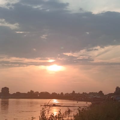 This is a photograph taken at sunset from the bank of a river. The sky is filled with clouds, and the setting sun casts a warm, orange glow over the scene. The river is calm, reflecting the colors of the sky. On the far bank of the river, there is a line of trees, and behind them, some buildings and parked vehicles are visible. The foreground of the image shows some vegetation along the riverbank. The location is in the village of Старый Буртюк, Russia.