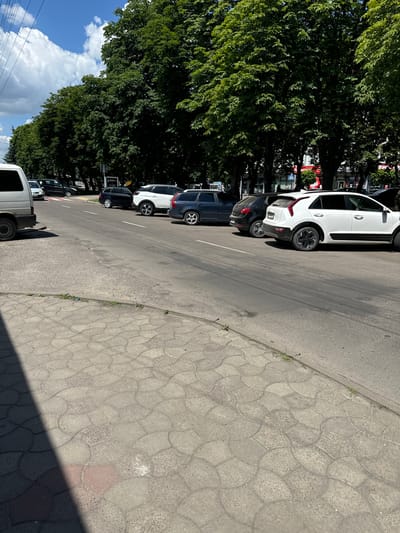 The image shows a row of parked cars along a street in Sarny, Ukraine. The cars are a mix of white and dark colors, and they are parked on the right side of the road, facing away from the camera. The street is paved with asphalt, and there is a sidewalk made of interlocking concrete pavers on the right. Large, leafy trees line the left side of the street, casting shadows on the road and sidewalk. The sky is blue with some fluffy white clouds. The overall impression is of a bright, sunny day in a suburban or semi-urban area. There are no people visible in the image. The activity appears to be simple parking.
Notable details include the distinctive pattern of the interlocking concrete pavers on the sidewalk, which are a common sight in many European countries. The trees are full and green, suggesting it is late spring or summer. The presence of power lines in the lower left corner indicates an urban or suburban setting. There is no visible text in the image.