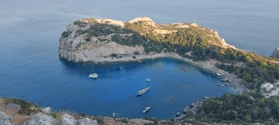 The image is an aerial, high-angle shot of a coastal bay, likely located near Faliraki, Greece. The bay is characterized by clear, bright blue water, with visible rocky outcrops and a sandy beach lined with numerous lounge chairs and umbrellas. Several boats are scattered across the water, including a large catamaran, a sailboat, and smaller motorboats. The surrounding land is rugged and mountainous, covered in lush green vegetation and trees, with steep, rocky cliffs. In the distance, the sea stretches out to a hazy horizon. The scene appears to be bathed in the warm light of either early morning or late afternoon, casting shadows across the landscape. A few small structures, possibly buildings or restaurants, are visible on the coastline near the beach. The overall impression is one of a serene and picturesque coastal resort.