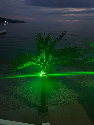 Nighttime scene at Shirokaya Balka, Russia. A palm tree is illuminated by a green light. The Black Sea is in the background under a cloudy sky at twilight. A pier is visible on the left side of the image, along with lights along the coastline. The ground around the palm tree appears to be covered in small stones. There is a wooden deck at the base of the palm tree.