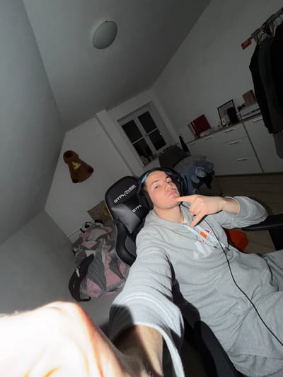 A young man, likely in his late teens or early twenties, is captured in a selfie-style photograph, holding the camera himself. He is positioned in a casual, slightly reclined posture, possibly in a chair, with his body angled. He wears a light grey hooded sweatshirt with a white and orange logo (possibly Ellesse) on the chest and has a black cable, likely from his headphones, across his chest. Around his neck or resting on his head are black headphones with "GTLPLAYER" and "GTP" visible on them.

The man's expression is playful, with a slight pout on his lips, and he points his right index finger towards his chin. His hair is short, and he has light stubble.

The setting appears to be an indoor room, most likely a bedroom, in Benningen, Germany. The walls are white. The room is dimly lit, suggesting it is evening or night, with artificial light casting shadows. In the background, to the left, a white window frame with dark panes is visible. Above the window, there's a circular ceiling light fixture. To the right, white cabinets or a wardrobe can be seen, along with shelves containing various items and dark clothing hanging. A brown stuffed animal, resembling a bear with a pizza slice design on its belly, is attached to the wall. Part of a patterned blanket or bedding (pink, grey, and white geometric design) is also visible, along with a partial view of another small stuffed animal, possibly a panda. The overall atmosphere is relaxed and personal.