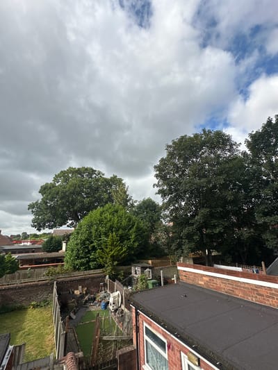 The image depicts an overcast sky above a residential backyard in Wrexham, United Kingdom. The foreground shows the rooftop of a building with dark grey roofing material. Beyond this, a brick wall separates the yard, which contains a patch of green grass, artificial turf, a garden shed, and various garden items including a white plastic chair and a blue bag. Several trees of varying sizes are visible, with some casting shadows on the ground. In the distance, residential buildings and more trees are present. The sky is filled with heavy, grey clouds, with patches of blue sky visible in the upper right quadrant. No people are present in the image.