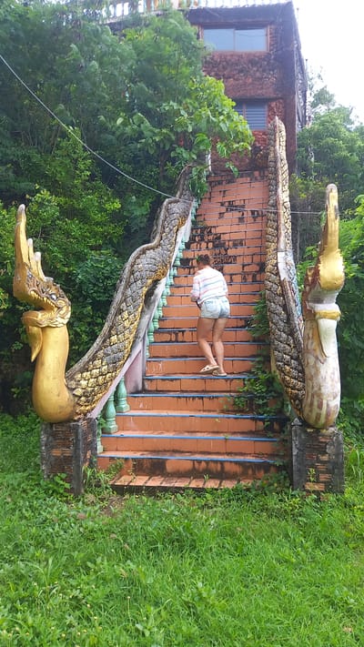 A medium-wide shot shows a woman on an outdoor staircase flanked by ornate dragon sculptures in Thap Put, Thailand. The staircase is made of orange brick with blue vertical bars between each stair. The dragon sculptures are gold and have a scale pattern. The woman is wearing a striped shirt and denim shorts. Lush greenery surrounds the staircase, including trees and grass. A building with a stone facade is visible in the background.