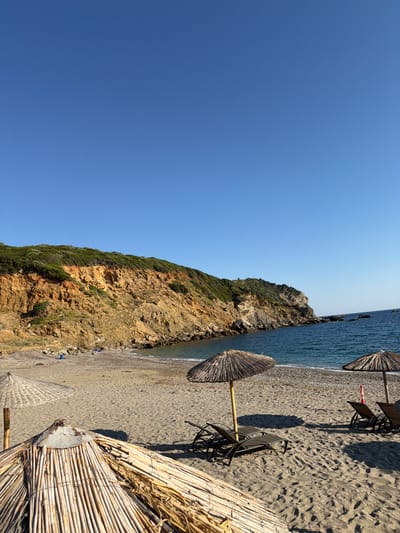 This is a photograph of a beach in Kalyvia, Greece. The scene is dominated by a large, rocky cliff face covered in sparse green vegetation, which rises from the shoreline. To the right of the cliff is the Aegean Sea, its water appearing calm and a deep blue. The beach itself is composed of dark sand and small pebbles.
Several beach chairs and umbrellas are set up on the sand, suggesting a leisurely day. The umbrellas are made of natural materials like straw and bamboo, typical of a Mediterranean beach setting. The chairs are dark and metallic. Shadows from the umbrellas and chairs are cast across the sand, indicating it is likely midday or early afternoon with the sun high in the sky. The weather appears clear and sunny, with a vibrant blue sky taking up a significant portion of the left side of the frame.
There are no people clearly visible in the foreground or mid-ground, though a few distant figures might be present along the shoreline or near the water. The overall mood is serene and inviting, characteristic of a relaxing beach vacation. The composition of the image, with the cliff and sea on one side and the beach amenities on the other, creates a balanced view of the natural landscape and the human-provided comforts. No visible text is present in the image.