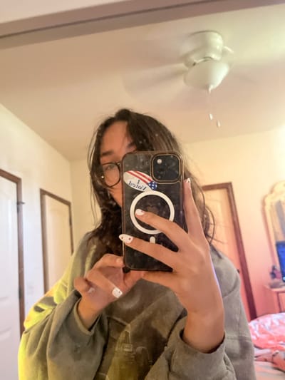 A female individual is depicted in a mirror selfie taken within a residential interior in Choctaw, United States. The subject has long, dark hair, wears eyeglasses, and is dressed in a grey long-sleeved garment. Her right hand, visible in the reflection, holds a dark-cased smartphone featuring a white circular MagSafe ring. An "I VOTED" sticker, displaying an American flag motif, is affixed to the phone case, appearing reversed due to the mirror. The nails on the visible hand are painted white with small red heart decals. A lighter skin patch is present on the visible forearm. The reflective background includes light-colored walls, a white ceiling with a stationary white ceiling fan/light fixture, and two interior doors. A decorative mirror frame and a source of blue/pink illumination, possibly from an electronic device, are partially visible on the right, adjacent to patterned fabric suggesting bedding.