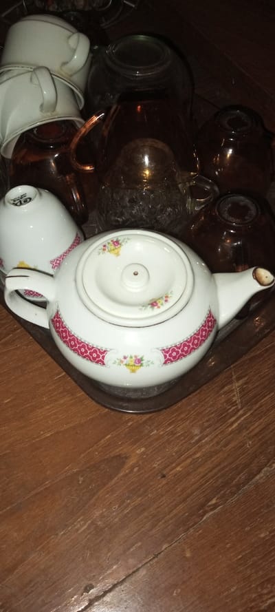 The image captures a collection of tea-serving items arranged on a tray, which rests on a worn wooden surface.
In the foreground and center, a white ceramic teapot with a lid is prominently displayed. The teapot features a distinctive design: a wide, fuchsia-pink band with a repeating patterned motif, accentuated by small floral basket designs on either side. Its lid also bears a similar delicate floral pattern. The teapot's spout shows some discoloration.
To the left of the teapot, a white ceramic teacup is visible, exhibiting a matching pink patterned band and floral accents. Behind and partially obscured, several other white teacups are stacked, some with gold-rimmed edges. One upside-down teacup in the mid-ground reveals text on its base, including "G&S" and potentially "CHINA."
The background features several dark brown glass items, including pitchers or decanters and smaller cups, which appear to be stored or part of a larger set. These glass items reflect ambient light, adding depth to the scene. All the items are situated on a dark, possibly metallic or plastic, tray. The overall lighting is somewhat dim, suggesting an indoor setting with low or artificial light. The wooden surface beneath the tray displays clear grain and signs of wear.
The scene depicts a still life arrangement of domestic tea ware, located in Aral, Kyrgyzstan.