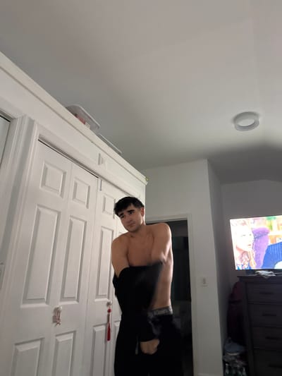 A young man is partially undressed in a room with white double doors in the foreground and a dresser with a television on top in the background. The man is facing left, pulling a dark garment over his head. He is wearing black underwear with "Calvin Klein" visible. The room appears to be a bedroom, with a light fixture on the ceiling. A doorway is visible to the right of the man. The television screen shows an image of two people. A white container with red accents is on top of the double doors. There is a red tassel hanging from one of the door handles. The location is Bellmore, United States of America.