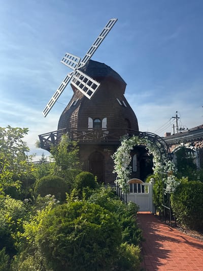 The image depicts a charming, two-story wooden building designed to resemble a windmill, situated in the city of Seltso, Russia. The building has a brown, shingled roof and a wooden balcony on the second level. It is surrounded by lush greenery, including various trees and bushes, some of which are meticulously shaped. A brick pathway leads to a white gate, which is adorned with an archway covered in white flowers. The sky is clear and blue with a few wispy clouds. The scene appears to be set in a garden or park-like environment, possibly a restaurant or a private residence, given the decorative elements and landscaping. There are no people visible in the image.