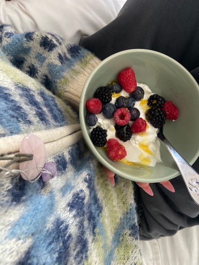 A close-up, medium shot shows a person seated, wearing a fuzzy, patterned sweater with shades of blue, white, and green. A bowl of yogurt with mixed berries (raspberries, blueberries, and blackberries) and drizzled with honey is held in front of them. A spoon is in the bowl. A necklace with a pink quartz pendant is visible on the sweater. The setting appears to be indoors, possibly on a bed. The specific location in the city of Welwyn Hatfield, United Kingdom, cannot be determined from this image.