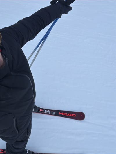 A first-person perspective shot of a person skiing. The individual wears a black jacket and gloves, holding blue and silver ski poles. The skier stands on a snowy surface with "HEAD" visible on the red skis. The background is primarily white snow. The angle is slightly overhead, focusing on the skier's upper body and skis.