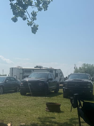 In Lyalta, Canada, on a bright, clear day, a residential scene unfolds with multiple vehicles and recreational equipment parked on a grassy area. A dark-colored sedan is parked on the left, partially obscured by trees. To its right, a black Ford F-150 pickup truck is prominent, with a silver and black travel trailer situated behind it. Further to the right, a black Chevrolet Silverado pickup truck is visible. The travel trailer appears to be a modern unit with a ladder attached to its rear. The ground is covered in green grass, with a few scattered white flowers. In the foreground, a dark brown, circular object, possibly a fire pit or planter, sits on the grass. A black folding chair, with a small black bag attached, is positioned to the right of the frame. Overhead, a branch with green leaves hangs from a tree, casting dappled shadows on the scene. The sky is a clear blue with a few wispy white clouds in the distance. There are no people visible in the image. The overall impression is of a recreational setting, likely a campground or a backyard during an outdoor gathering.