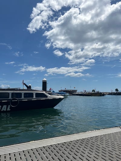 This image depicts a sunny day at a harbor in the city of Tidore Islands, Indonesia, with a clear blue sky dotted with fluffy white clouds. The foreground features a section of a gray, textured dock, with the calm, dark blue water of the harbor stretching out before it. In the midground, a sleek, dark-colored passenger boat is docked, with windows along its side and the partial word "GAL" visible on its hull. A spare tire hangs from its side. Several other boats are moored further in the harbor, some with flags, and a long structure that appears to be a building or pier with a roof is visible on the shore in the distance. The scene is serene and suggests a typical day at a coastal Indonesian port.