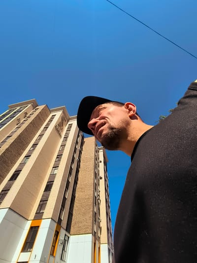 An exterior, eye-level, low-angle shot shows a man wearing a black baseball cap and a black shirt, looking up towards the sky. He has stubble and is smiling. To the left, a tall, modern apartment building with beige brick and white panels rises into the clear blue sky. The building features numerous windows and some orange accents. Power lines are visible against the sky. The scene is set in Karaganda, Kazakhstan.