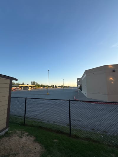 The image, when viewed upright, displays an outdoor scene encompassing a large commercial or industrial building, an extensive parking lot, and a chain-link fence under a clear sky. A substantial, light-colored building with a flat roof occupies the left side of the frame, showing architectural details consistent with a warehouse or large retail store. Its walls reflect warm light, suggesting a low sun angle.

A black chain-link fence stretches across the middle ground, acting as a boundary. To the right of the fence, there is a strip of green grass mixed with drier, lighter patches of earth. Beyond the fence, a wide, empty asphalt parking lot extends into the distance, dotted with numerous tall light poles. Some red painted lines are visible on the asphalt closer to the building. In the bottom right foreground, the corner of a shed-like structure, featuring light-colored horizontal siding and a dark roof, is partially visible.

The sky is a bright, clear blue with a hint of haziness or very thin clouds in the upper left. The soft, long shadows cast by the building and fence indicate either early morning or late afternoon/early evening. The parking lot appears deserted, with no vehicles or people present, contributing to a calm and quiet atmosphere. The scene suggests a commercial or industrial area within Anchorage, United States of America. There is no visible text in the image.