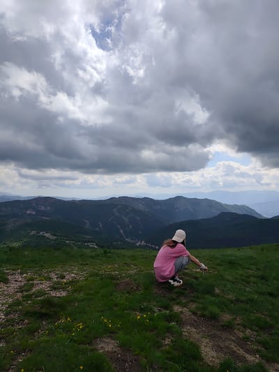 The image displays a mountainous landscape in Sedlačka gora, Montenegro, under a heavily clouded sky. In the foreground, a single person is seen from behind, crouching on a grassy slope. The individual wears a light-colored baseball cap, a pink short-sleeved t-shirt, grey pants, and sneakers with distinct black and white wavy soles. Their left arm is extended forward, with the hand appearing to point or interact with the ground. The immediate ground cover is green grass, dotted with small yellow wildflowers, and interspersed with patches of exposed soil and small rocks. Beyond the foreground, successive layers of dark green, tree-covered mountains extend into the distance, appearing progressively lighter and bluer due to atmospheric perspective. Visible clearings or structures are present on some of the mountain slopes. The sky is predominantly covered by thick, dark grey clouds, with a notable patch of blue sky and lighter clouds visible towards the upper center-right.