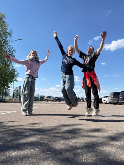 The image shows three people standing on top of each other in what appears to be a parking lot. The person at the bottom is wearing a black t-shirt, black pants, and white sneakers, with a red jacket tied around their waist. The middle person is wearing a navy blue sweatshirt and jeans. The person on top is wearing a light pink long-sleeved shirt and light-colored jeans, with a travel neck pillow. All three people have their arms outstretched. The background includes parked cars, a paved surface, trees, a building, and a blue sky with scattered clouds. The location appears to be Аэропорт Уфа, Russia.