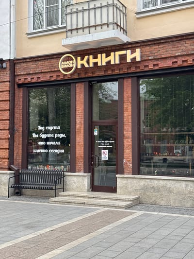 The image depicts the storefront of a bookstore in Akhieli, Georgia. The sign reads "Books Amon Bilon" vertically along the red brick facade of the building. The windows have a quote on them "A year from now you'll be glad you started today." A black bench is in front of the window. The entrance has a dark-framed glass door with signage including "No smoking". The storefront is adjacent to a sidewalk. The building is several stories high.
