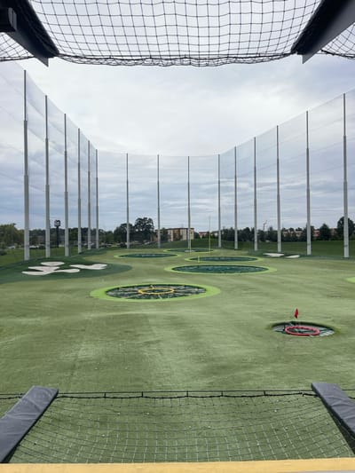 The image displays a golf driving range facility in Oxon Hill, United States, viewed from an elevated bay. In the immediate foreground, a black mesh safety net spans the width of the frame, with a yellow horizontal barrier directly beneath it and dark grey padded rails positioned on the left and right. The expansive playing field is covered in artificial green turf, featuring numerous circular targets of varying sizes. These targets include dark central areas, some encircled by distinct yellow or red rings. A small red flag marks one target hole on the right side of the field. White golf balls are scattered across the turf. Three irregularly shaped white patches, resembling sand bunkers, are situated on the left half of the range. A tall, yellow goalpost, consistent with American football goalposts, stands vertically towards the rear-left of the field. The entire driving range is enclosed by a high perimeter net supported by numerous light grey vertical poles that curve around the playing area. Beyond this enclosure, trees, several multi-story buildings, and a paved area with parked cars are visible in the distance. The sky above is uniformly overcast. No individuals are present in the scene.