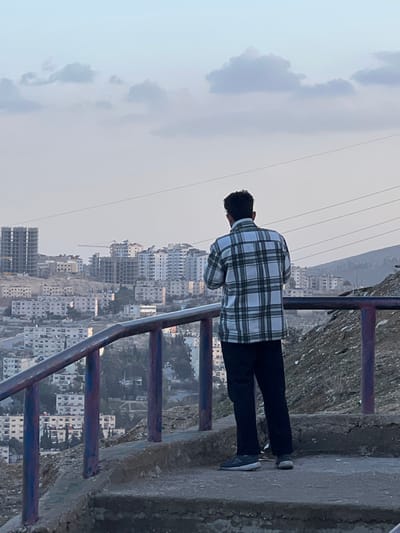 A male individual is depicted from behind, standing on an elevated concrete platform overlooking the city of Qudsaiya, Syria. He wears a white and green plaid shirt, dark trousers, and light-colored athletic shoes. His hands are positioned in front, holding an object directed towards the panoramic cityscape. A worn metal railing, painted with purple and blue sections, extends across the foreground. To the right, a rocky, sparsely vegetated hillside is visible. The urban landscape below features numerous multi-story residential buildings, some appearing completed and others under construction with visible concrete structures and cranes. Multiple power lines traverse the sky, which is partly cloudy. The lighting suggests late afternoon or early evening conditions.