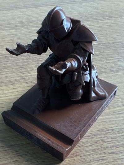 The image shows a 3D-printed figurine of a crouching figure wearing armor, possibly a knight or rogue, emerging from a rectangular object. The rectangular object seems to be a wall or a pedestal, with some ornamental detailing along its top edge. The entire scene is placed on a surface with a wood grain pattern. The lighting in the scene is diffused, with no prominent shadows. Based on the limited information, it is impossible to determine the exact location within Чыжэвіцкі сельскі Савет, Belarus.