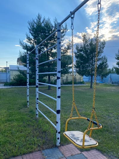 An outdoor scene in the city of Volzhsk, Russia, features a yellow swing set with a worn wooden seat, suspended by chains from a metal frame. The frame is painted white with black splotches. Adjacent to the swing set is a white metal structure with horizontal bars, also marked with black splotches, resembling playground equipment or an exercise bar. The ground is covered in green grass, with a paved walkway made of red and grey bricks in the foreground. In the background, several evergreen trees are visible, along with a light-colored building, a greenhouse, and a fence. The sky is partly cloudy with the sun breaking through, suggesting late afternoon or early evening light. There are no people in the image.