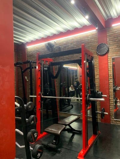 The image shows the interior of a gym in Kecamatan Kartasura, Indonesia, with a weightlifting machine in the foreground. The machine has red, black, and silver accents, and is set against a brick wall with a red and white accent stripe. There is a black bench with a brown leather seat and two sets of weights. The wall is mirrored on one side, reflecting a woman in a headscarf and a man in a black tank top sitting on a bench and looking at the camera. There is a large clock on the wall behind the machine. The ceiling is a corrugated metal structure with some fluorescent lights. The floor is black, and the room is well lit. The gym appears to be clean and well-maintained. There are other pieces of gym equipment visible in the reflection of the mirror.