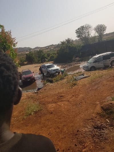 The image depicts an outdoor scene featuring a very muddy, unpaved dirt road. The photograph is oriented vertically, showing a bright, clear sky dominating the upper left portion.
In the middle ground, three vehicles are visible navigating or parked along the extremely challenging road. The road surface is a reddish-brown earth, indicative of laterite soil, and is heavily inundated with large, murky puddles.
The vehicle closest to the viewer, positioned on the right side of the road, is a white or light-colored van or minibus. It appears to be partially submerged in a substantial puddle, suggesting it might be stuck or proceeding with extreme caution. Faint, dark markings are visible on its side, but they are illegible due to the distance and resolution.
Behind the white van, a second white car, possibly a sedan or small SUV, is also situated within or near a large puddle, its surface reflecting the bright sky. Further back, and partially obscured by the second white car and another puddle, a red car can be seen. All three vehicles appear to be facing generally in the same direction, towards the left of the frame.
Lush green bushes and trees line both sides of the muddy road. On the left side, larger trees with distinct, somewhat sparse canopies extend towards the horizon. In the far distance, faint outlines of hills or elevated terrain are visible under the clear sky.
The sky is bright and appears to be a light blue or off-white, indicating fair weather. The bright illumination suggests it is daytime, likely late morning or early afternoon, given the overall lighting and lack of prominent long shadows.
In the immediate foreground, particularly in the lower right corner, a blurred, dark shape is visible, which appears to be the top of a person's head and shoulder, suggesting the photograph was taken from a first-person perspective at eye level or slightly looking down.
The scene conveys the difficulty of travel on poor infrastructure, with vehicles contending with significant mud and water obstacles on what seems to be a common thoroughfare in a semi-rural or developing area. There is no clearly legible text visible in the image.