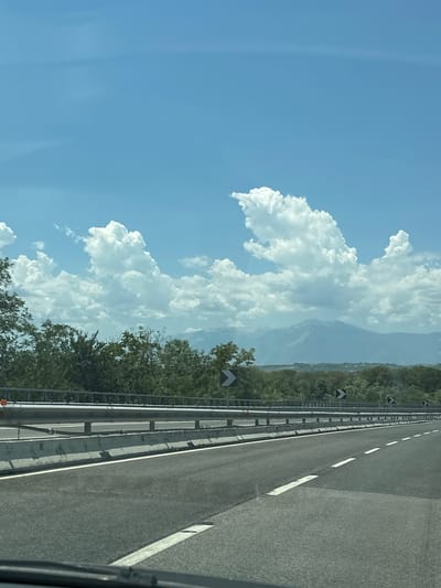 The image shows a highway with a concrete barrier and guardrail on the left side. The road is asphalt with white lane markings. There are trees and vegetation on either side of the highway, and mountains in the distance under a bright blue sky with fluffy white clouds. The scene appears to be a daytime shot taken from a moving vehicle, suggesting a travel or commute context. The location is identified as the city of Paglieta, Italy.