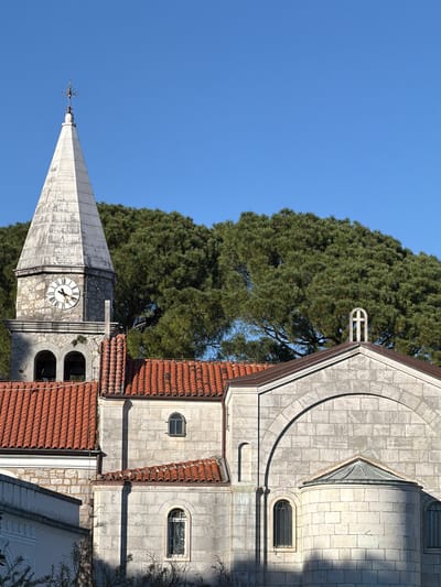 The image shows a building with a clock tower, likely a church or other religious structure, in the city of Grad Opatija, Croatia. The architectural style appears to be Mediterranean or Adriatic, with stone construction and red tile roofing. The clock tower has a conical spire. The sky is clear and blue. A large, dense tree stands behind the building. The image appears to be taken from a low-angle perspective.
