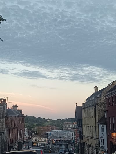 The image shows a street scene in Arundel, United Kingdom, likely during the late afternoon or early evening, with a pale pink and blue sky. The sky is filled with a textured pattern of grey and white clouds. In the foreground, a narrow street winds downhill, lined with historic brick and stone buildings. Some of these buildings appear to be shops, with signs visible, including one that reads "Tallant". Construction or renovation is indicated by scaffolding covering some of the buildings. Vehicles are present on the street, suggesting it is a functional thoroughfare. The background features rolling green hills covered in trees.