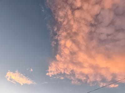 The image shows a sky with scattered clouds. The clouds on the right side of the frame are illuminated with warm, rosy hues, suggesting either sunrise or sunset. The sky on the left is a clear, pale blue. A thin, dark wire stretches diagonally across the lower right portion of the image. There are no people or specific landmarks visible that would indicate a location within Konstantinovka, Russia. The scene is a natural one, depicting atmospheric conditions.