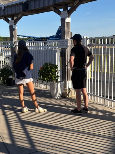 In the Town of East Hampton, United States of America, a young woman and a young man stand on a paved walkway outside a small airport. The woman, facing away from the camera, wears a navy blue t-shirt, a white skirt, a navy blue baseball cap, and white sneakers. She has a navy blue backpack slung over her shoulders. The man, also facing away, is dressed in a black short-sleeved shirt, black shorts, a black baseball cap, and black sneakers. Both individuals appear to be waiting.
Behind them, a white fence runs along the perimeter of the airport tarmac. A small propeller airplane with "KODIAK" visible on its tail is parked on the tarmac. The environment is sunny with distinct shadows cast by the fence on the ground. Two potted plants, one with green foliage and the other with a red flower, are placed near the fence. A sign on the fence reads "Passengers And Crew Only Beyond This Point." Another sign on the fence further down reads "PUSH." The scene suggests a departure or arrival at a small regional airport on a clear day.