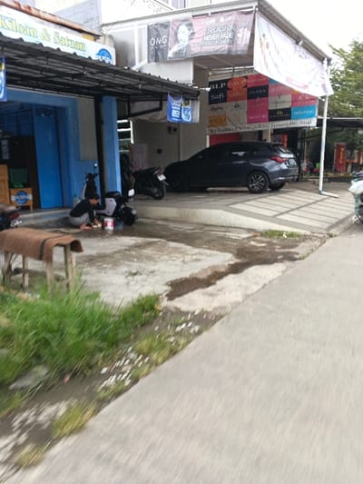 This image captures an urban street scene in Rancamanyar, Indonesia. The foreground displays a paved road on the right, transitioning to a narrower concrete path. To the left of this path, a strip of green grass and damp dirt is visible, along with a partial view of a wooden bench topped with a brown mat.

In the midground, a wet concrete surface extends, serving as a driveway or service area. On the left side of this area, in front of a blue-painted storefront, a person is crouched. They wear a dark long-sleeved top and light pants, and appear to be cleaning a light-colored motorcycle or the ground with a white bucket beside them. Behind this activity, another dark motorcycle is parked. The blue storefront features a dark metal awning and signs advertising "Kiloan & Satuan" and "Puspa."

To the right, a wider section of the building has white walls, surmounted by a large white awning. This awning supports several advertising banners for cosmetic brands (including OMG, BLP, Safi, Salsabila, YOU, Reglow, Dazzle Me, Skintific), some with Indonesian text like "Selalu On Never Fade Matte Kiss Lip Cream." Beneath this awning, a dark-colored SUV is parked, facing right. A partially visible motorcycle is also present on the far right edge of the frame.