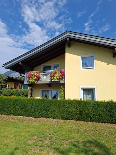 The image shows a yellow house with a balcony and flowers in Münster, Austria. The house has a dark roof with exposed wooden beams. The balcony has a railing and is decorated with hanging baskets of pink and yellow flowers. There are two windows visible on the side of the house, both with white frames. A dense green hedge runs along the front of the property, separating it from a grassy lawn. In the background, a mountain can be seen under a blue sky with white clouds. To the left, another building with a wooden facade is partially visible. The scene is brightly lit, suggesting it is daytime and sunny. There are no people visible in the image.