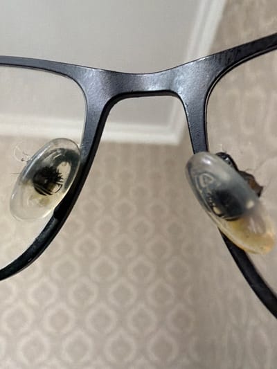 A close-up, eye-level, interior shot shows the nose pads of a pair of black-rimmed glasses. The nose pads are clear, oval-shaped, and appear to be made of plastic or silicone. They are attached to the frame by thin metal arms. The left nose pad has some debris and lint stuck to its surface, and the right nose pad appears discolored and dirty. The background is a light-colored wall with a subtle geometric pattern. The lighting is soft and diffused, suggesting an indoor setting. The image does not provide any information about a specific city or location in Belarus.