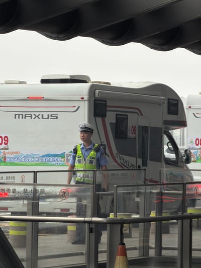 A uniformed Chinese police officer stands near a white recreational vehicle (RV) with "MAXUS" written on its side. The officer is wearing a bright yellow vest with "JING CHA" (police) written on the back in Chinese characters. The RV has a license plate that appears to be red and white. In front of the RV, a transparent barrier displays Chinese text and symbols, including a red circle with a diagonal line through it, indicating a prohibition. Yellow and grey bollards are visible behind the barrier. The scene appears to be in an outdoor, possibly a city, environment, given the presence of the police officer and the context of the vehicle markings and signage. The lighting suggests it is daytime.