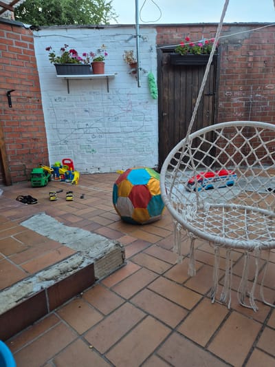 The image depicts an outdoor patio area, likely a backyard, with terracotta tiled flooring. A white macrame hanging chair is prominently featured in the foreground, to the right. In the background, a brick wall on the left has various drawings and scribbles. A white brick wall with mounted planters containing red and pink flowers is visible. A wooden door is set into a brick wall on the right. Scattered across the patio are children's toys, including a green toy truck, a yellow and blue toy construction set, and a large, multicolored fabric ball. There are also a pair of black sandals and some yellow children's shoes. The scene appears to be in the city of Aalst, Belgium.