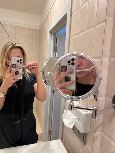 A woman with blonde hair is taking a selfie in a bathroom mirror. She is holding her phone in her right hand and using a white stick with the other hand. A smaller mirror is on the left. The mirror reflects her face with the white stick near her lip. The bathroom has tiled walls, a tissue dispenser, and a sink. She has a watch on her left wrist. The phone case has stickers on it.