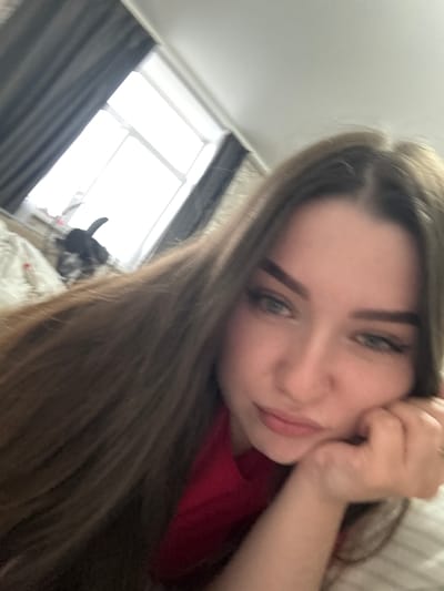 A slightly tilted, close-up indoor shot features a young woman in Savenki, Russia. She is the primary subject, looking towards the camera with a gentle, relaxed expression. Her long, straight light-brown hair frames her face, and her light-colored eyes are clearly visible. She wears a red top, and her hand is resting near her temple or cheek. In the blurry background, a window with bright light filtering through suggests it is daytime. Dark curtains partially cover the window. The setting appears to be a room, possibly a bedroom or living area. No text is visible in the image.